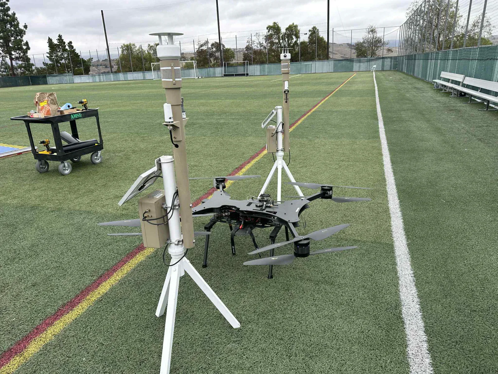Two ground detection masts on tripods beside a black multirotor aircraft parked on a sports field