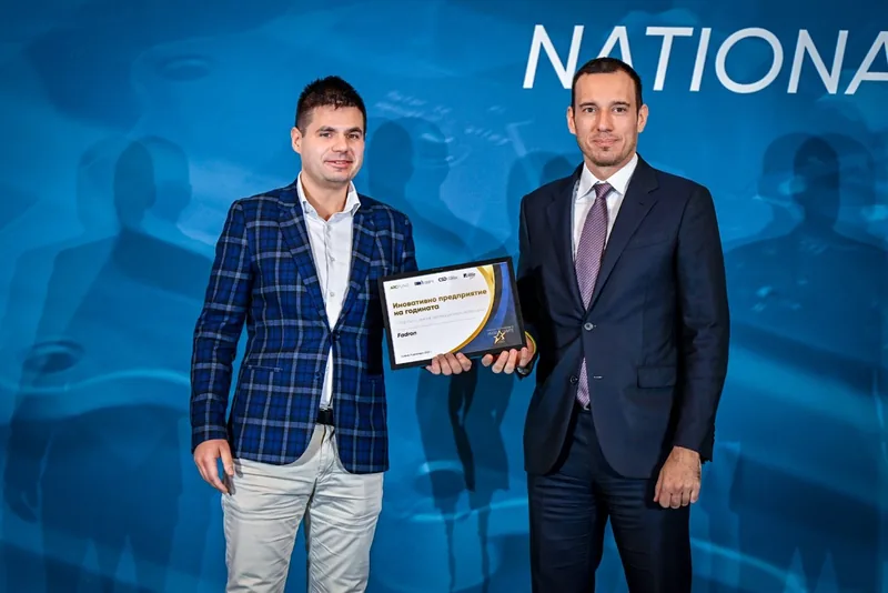 Two men on a blue-lit stage holding an award certificate at the National Innovation Forum